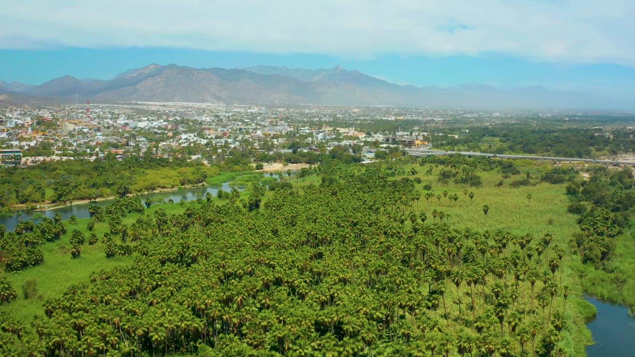 At 0:02, green palms and fields fill the lower frame, a narrow river channel sits left of center, and the San Jose del Cabo town and mountain range remain visible, with no visible people.