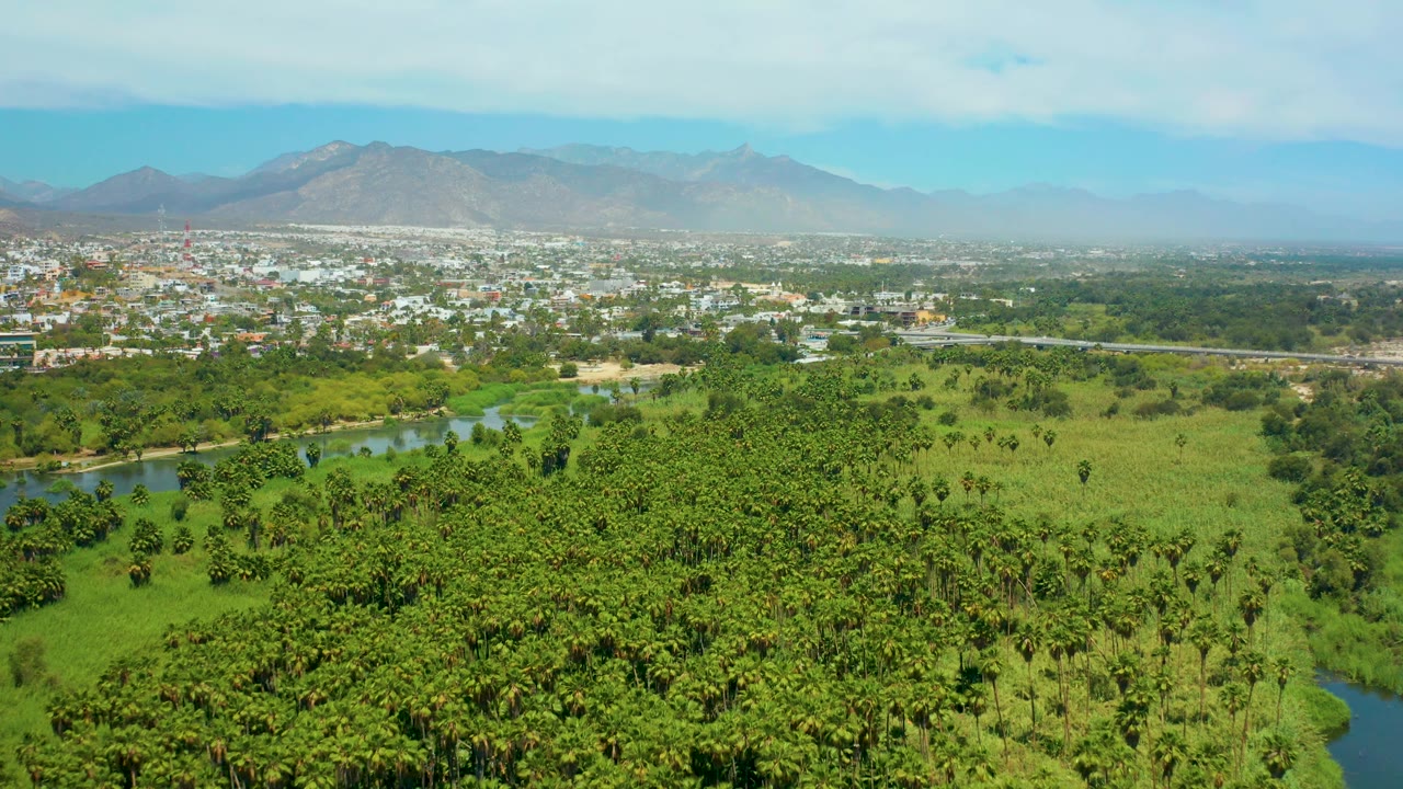 Aerial countryside view with river, green fields, low buildings, and mountains.