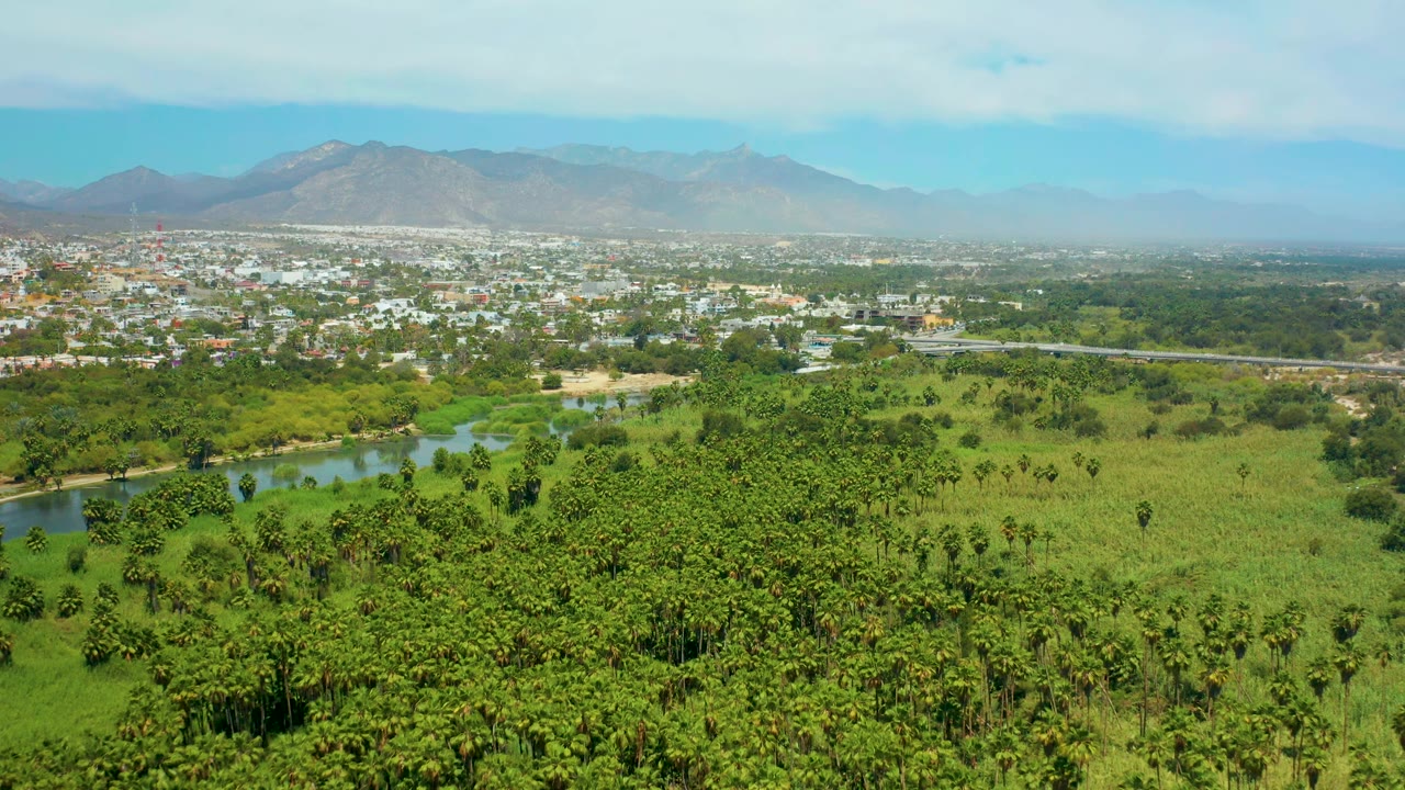 Second aerial frame of San Jose del Cabo showing the river bend partly framed by trees, broad green fields, low buildings in the distance, mountains, pale clouds, and no visible people.