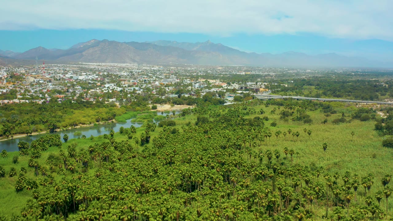 Third aerial frame in San Jose del Cabo with a green field pattern across the foreground, river water left of center, distant town structures, mountain ridge, and no visible people.