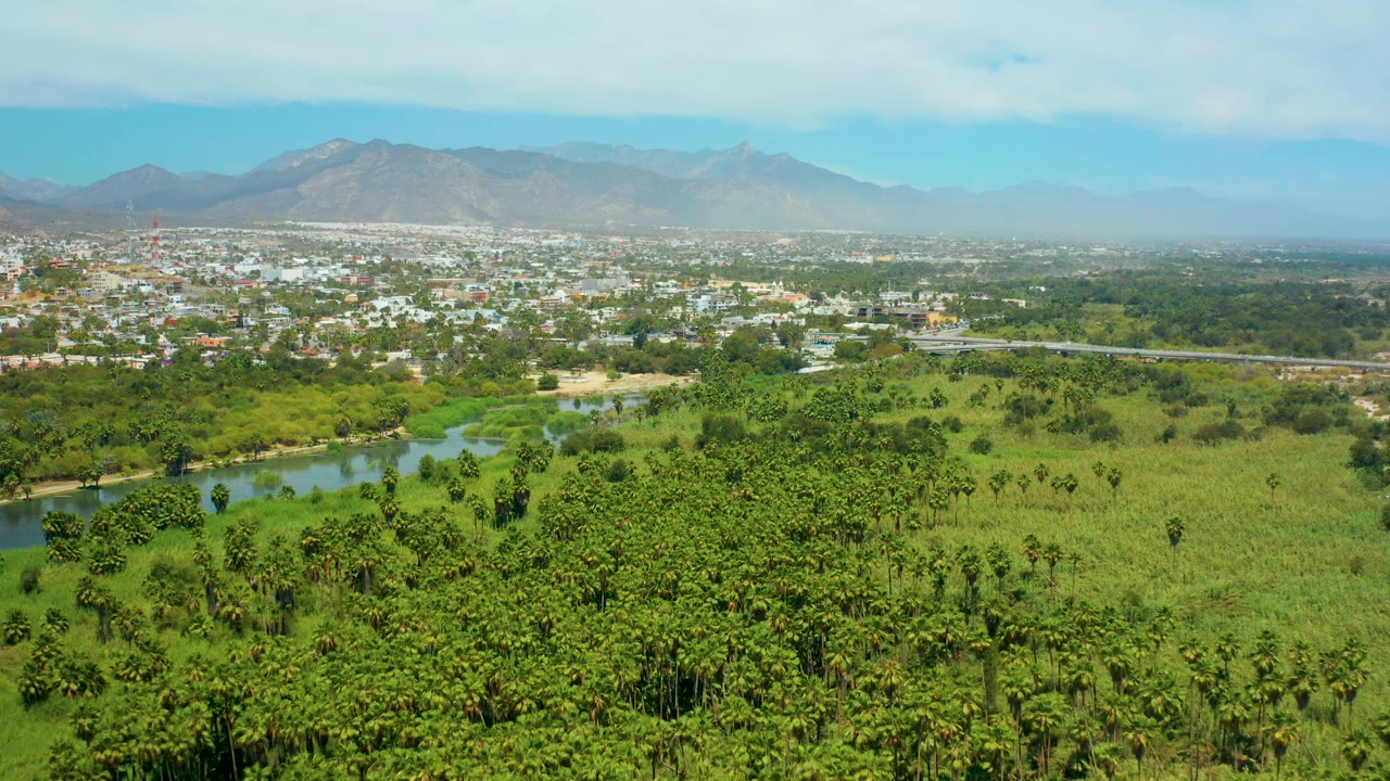 Aerial frame with river channel, fields, scattered trees, town, and mountains.