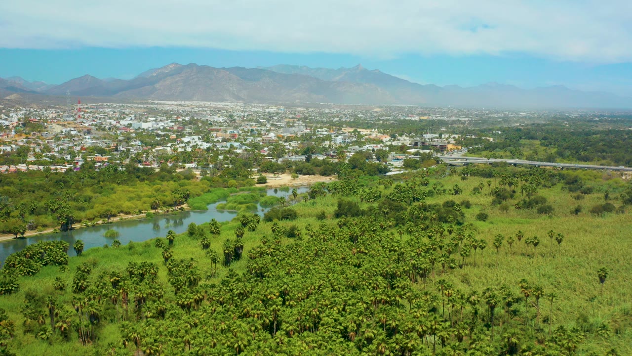 Opening aerial frame over San Jose del Cabo shows a river bend, dense green vegetation, town buildings, hazy mountains, blue sky, and no visible people.