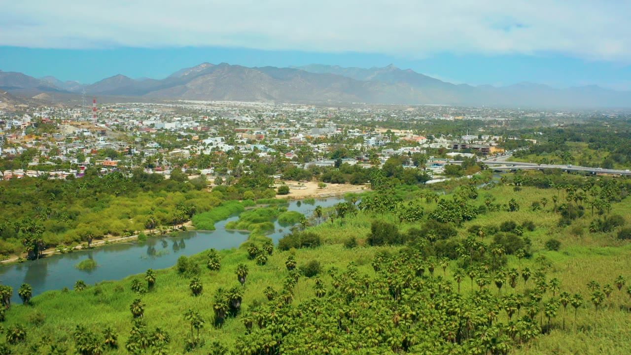 Wide aerial view over San Jose Del Cabo with a winding river in green fields, a spread of town buildings, hazy mountains, partly cloudy sky, and no visible people.