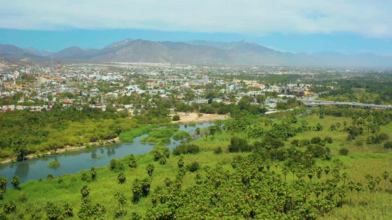 Aerial frame of San Jose Del Cabo with the river curving through dense green vegetation, white town buildings beyond it, distant mountains, and no visible people.