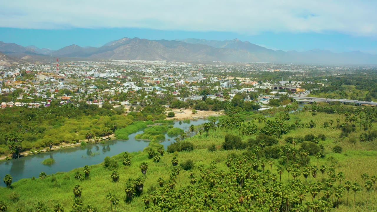 High aerial view in San Jose Del Cabo showing blue-green water, palms and brush in the foreground, a low urban area, mountains, thin cloud cover, and no visible people.