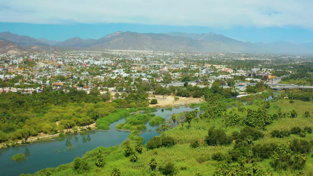 San Jose del Cabo landscape with the river curve more centered below thick green vegetation, white and tan town buildings across the middle distance, hazy mountains, and no visible people.