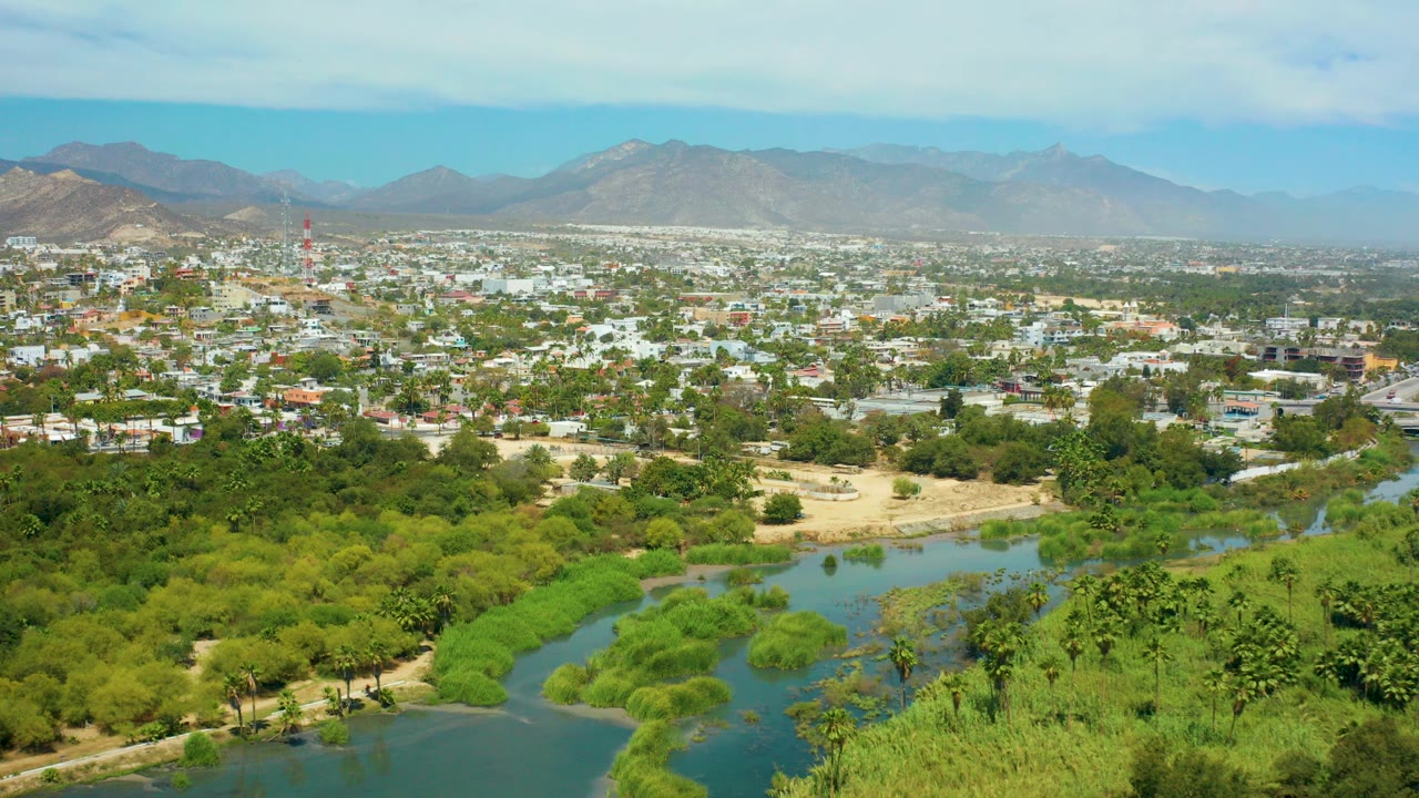 Wide aerial view over San Jose del Cabo with green river water and trees in the foreground, low city buildings beyond, distant mountains, and no visible people.