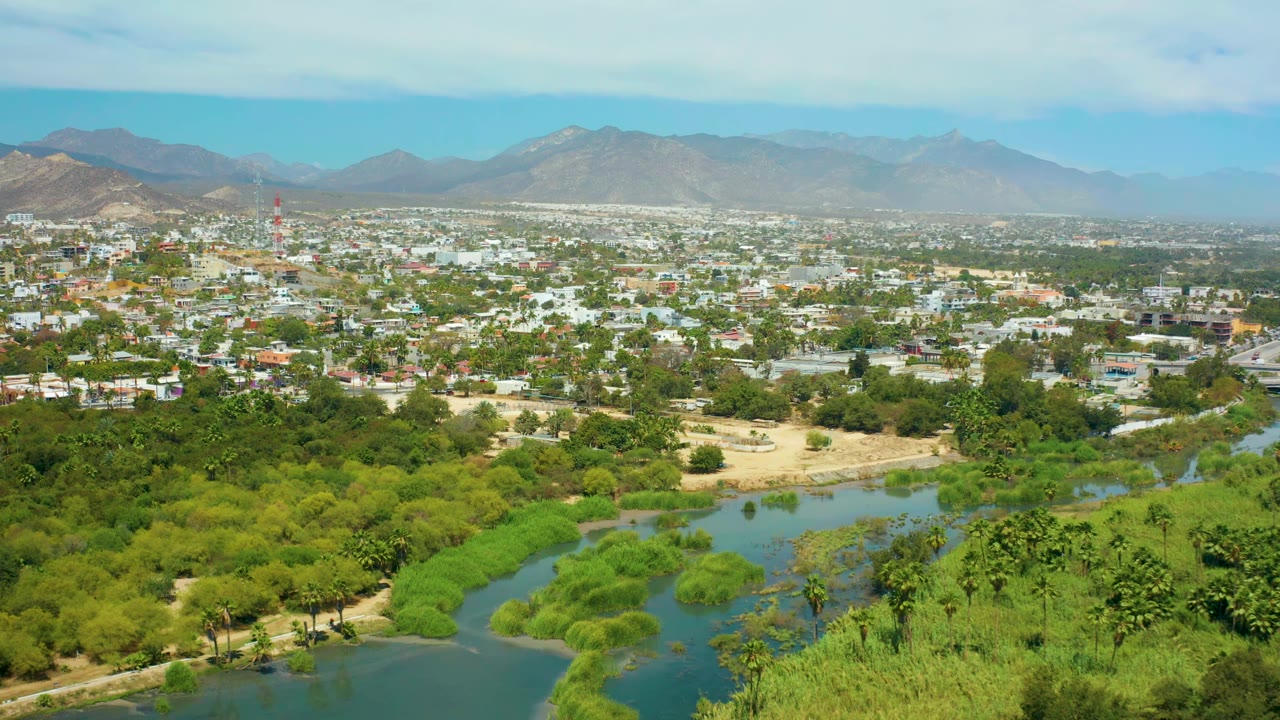 High aerial frame in San Jose del Cabo showing clustered green vegetation around the river, pale city blocks across the midground, hazy mountains, and no visible people.