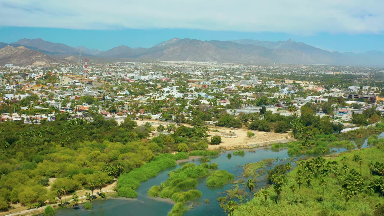 Aerial view over San Jose del Cabo with bright green river vegetation in front, compact urban development in the distance, mountains on the horizon, and no visible people.