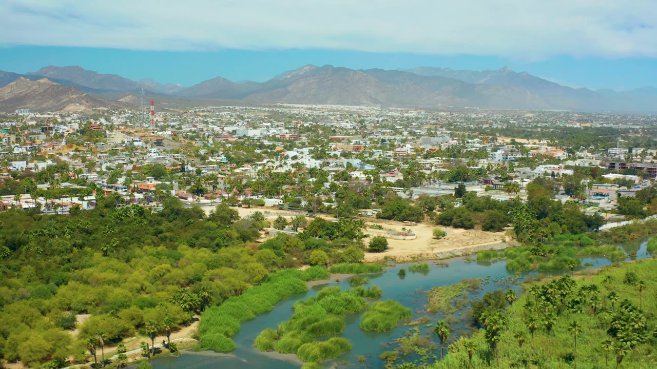 Wide aerial of green river bends below an urban area and mountains.