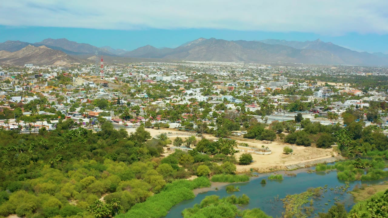 Opening aerial frame over San Jose del Cabo, with a green river corridor at the bottom, dense low-rise city blocks beyond it, distant mountains under pale cloud cover, and no visible people.