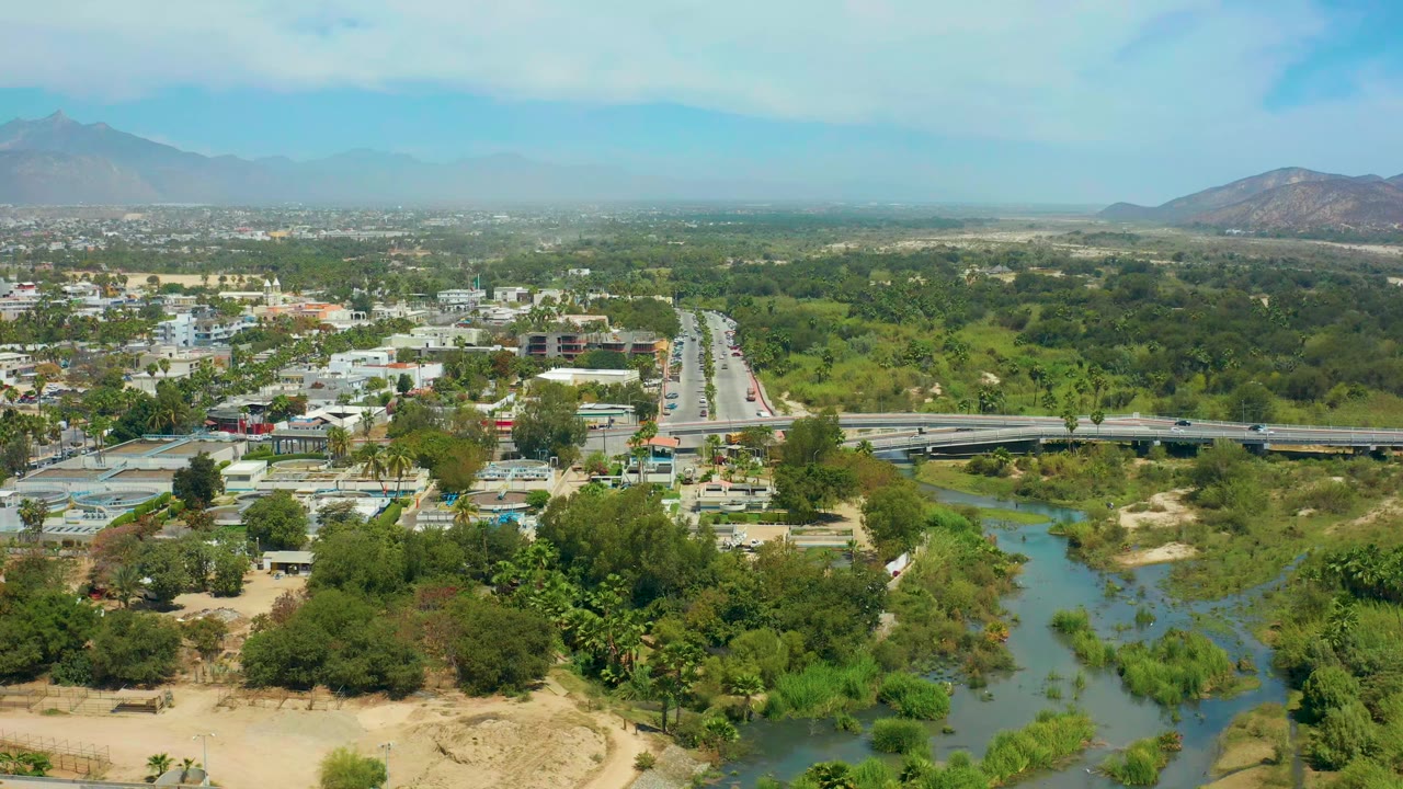 Wide San Jose Del Cabo aerial with the river corridor centered below, the road and bridge crossing the midground, scattered buildings among trees, hazy mountains, and no visible people.