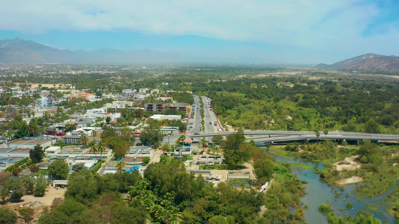 Opening aerial frame over San Jose Del Cabo with a divided road leading toward a bridge, green river corridor, low-rise buildings, distant mountains, scattered cloud, and no visible people.