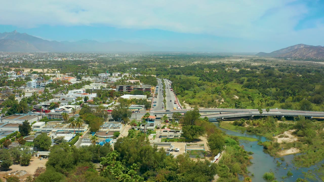 At 0:02, the drone perspective shows the bridge across the lower frame, river water and sandy banks below, traffic on the central road, San Jose Del Cabo buildings, hazy mountains, and no visible people.