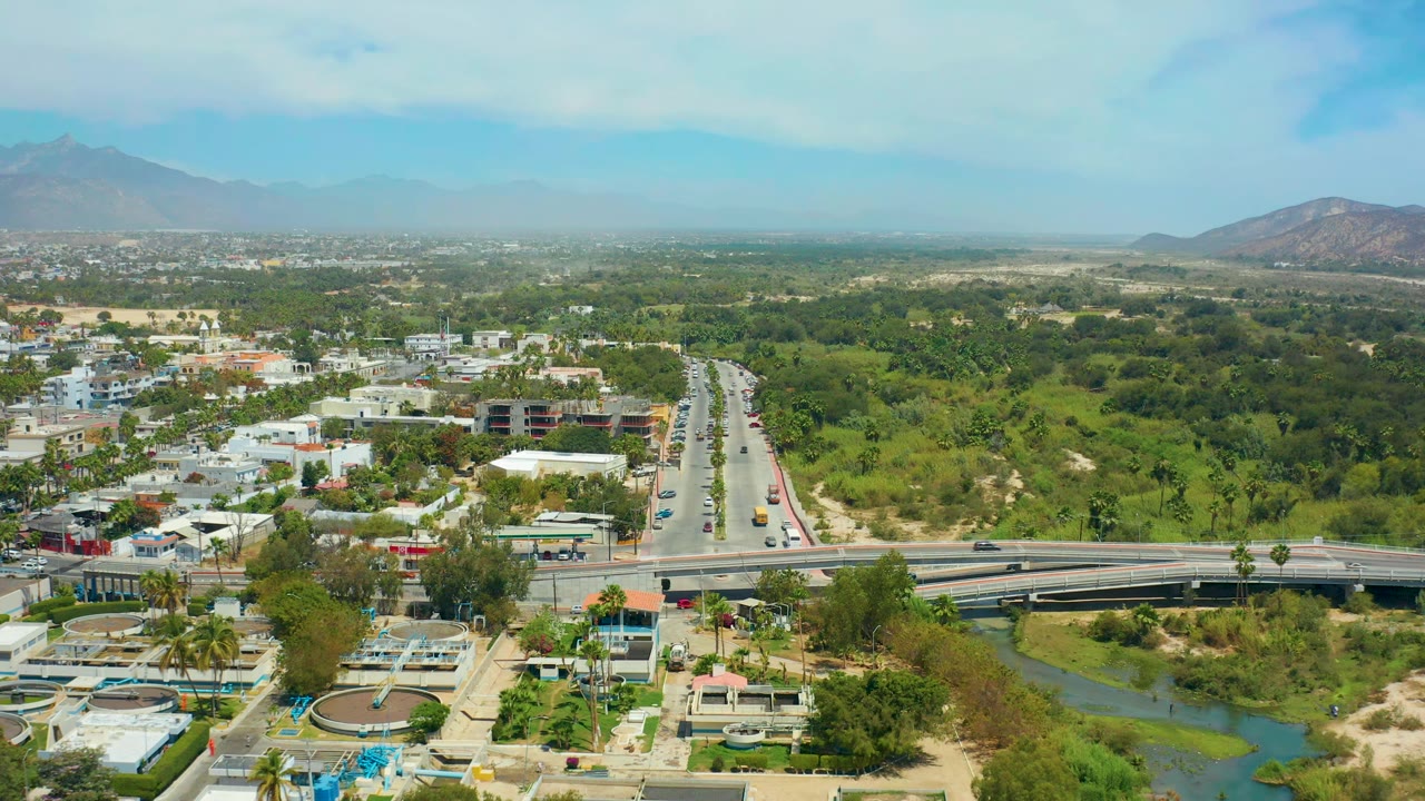 One second into the clip, the San Jose Del Cabo aerial view centers the straight roadway running toward the horizon, with the bridge and river channel below and no visible people.