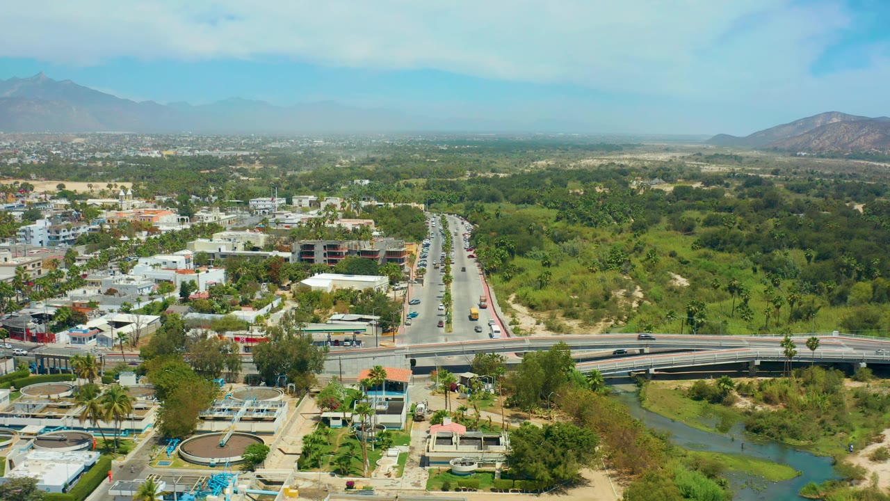 At 0:02, San Jose Del Cabo appears from above with vehicles on the highway, a curved bridge in the foreground, dense green vegetation to the right, hazy mountains, and no visible people.