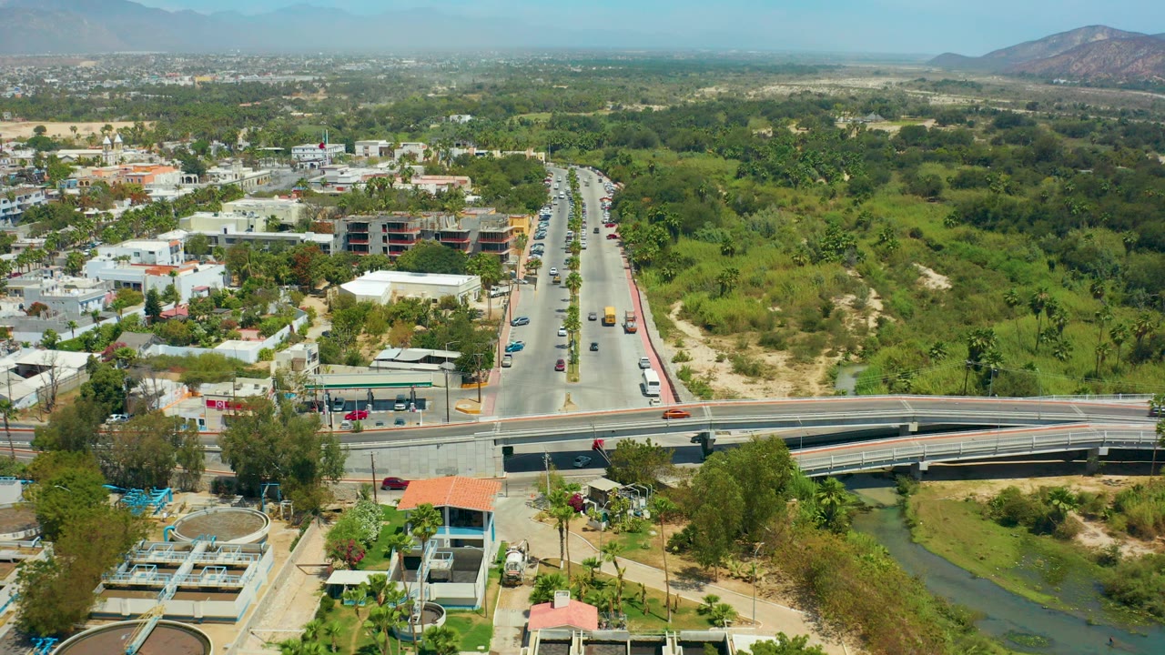 Aerial daytime view in San Jose Del Cabo, Baja California Sur, with a divided road running toward distant hills, an overpass and canal in the foreground, scattered cars, low buildings, greenery, and no visible people.