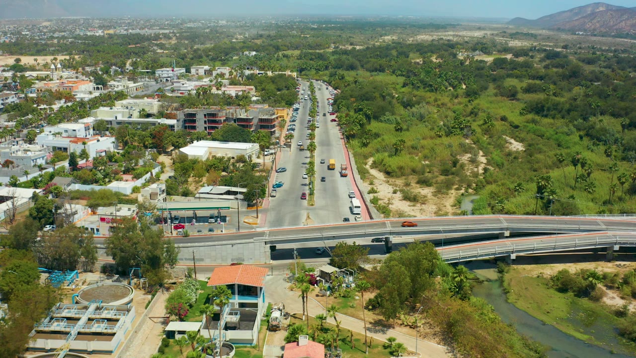 Drone view of traffic on a divided road beside greenery and low buildings.