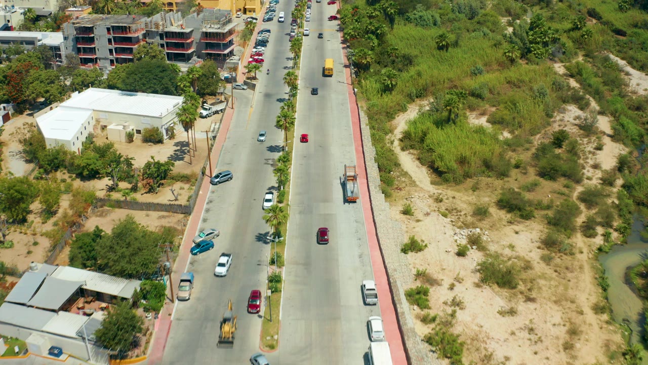 Aerial frame from San Jose del Cabo showing a multi-lane road with scattered cars, a white building, construction structures, dry vegetation, and no visible people.