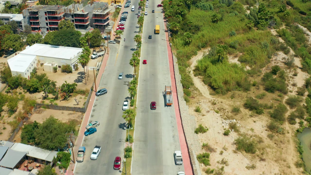 San Jose del Cabo aerial frame with the road centered, red and white vehicles in traffic, a sidewalk, a large green and sandy lot, low buildings, and no visible people.