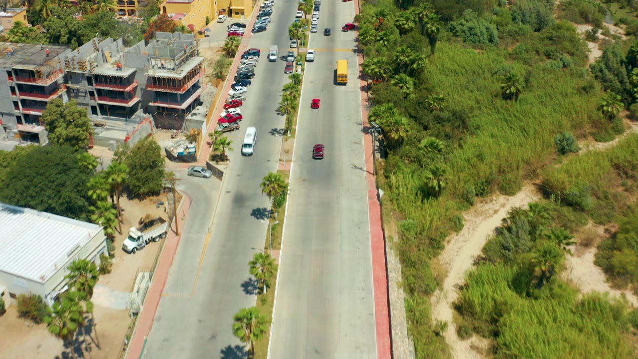 Overhead view in San Jose Del Cabo showing a paved road, a concrete building under construction, parked cars along the curb, dense greenery, and no visible people.