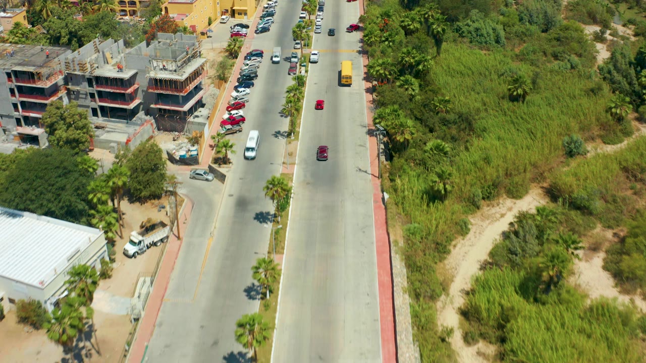 Aerial frame in San Jose Del Cabo with a yellow bus and cars on the road, parked vehicles beside yellow buildings, a construction site, roadside greenery, and no visible people.