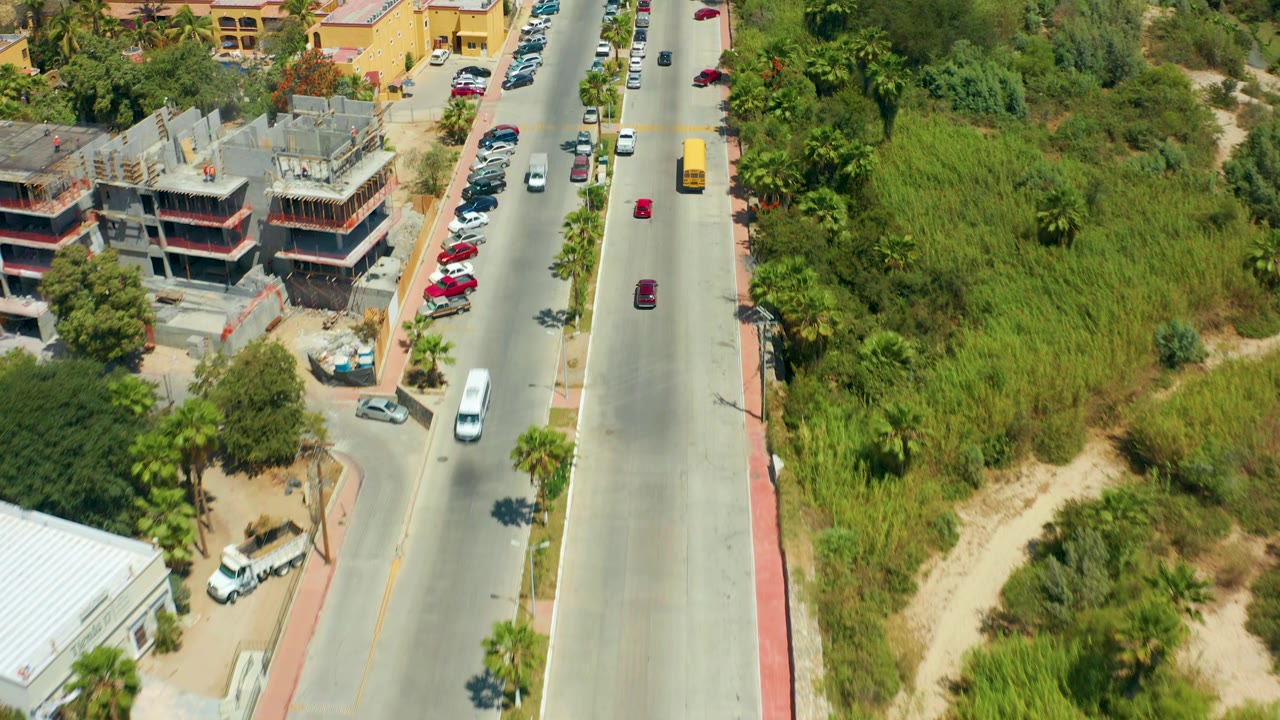 Overhead San Jose Del Cabo road scene with multiple cars, a yellow bus farther up the lane, parked cars, a partially built concrete structure, scrubby green lots, and no visible people.
