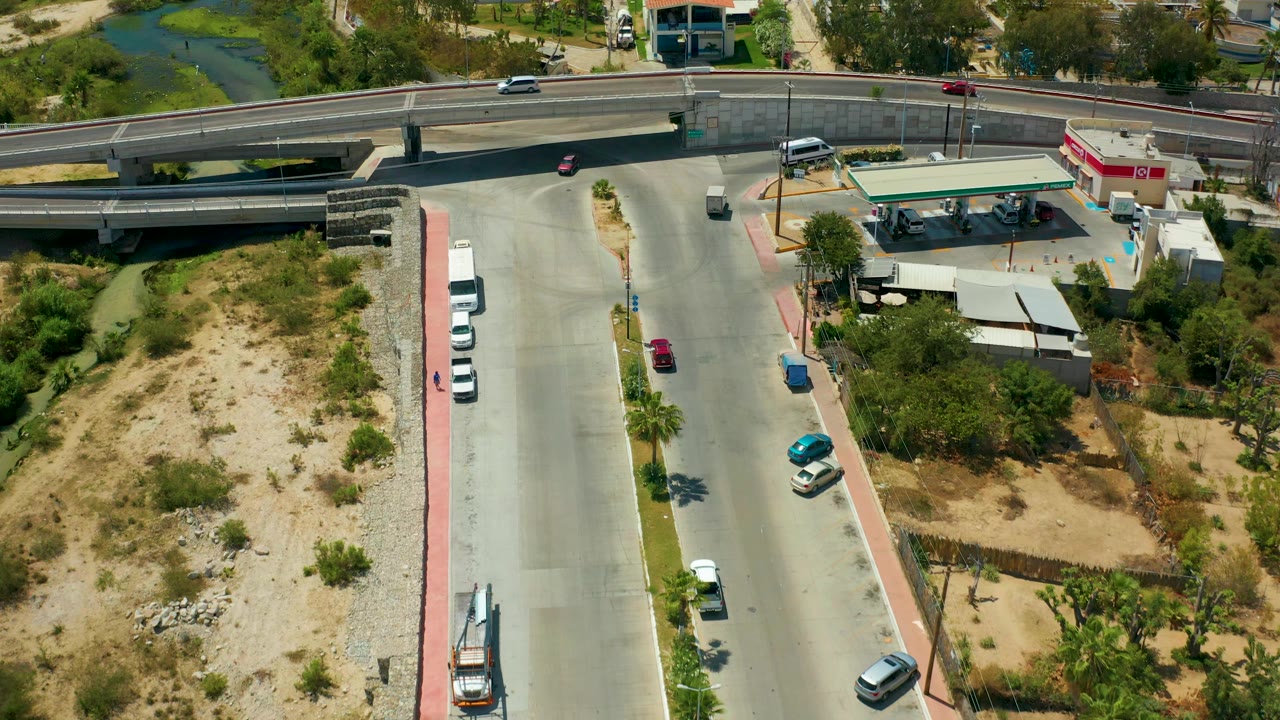 San Jose del Cabo aerial frame with cars on the road, the overpass crossing the upper view, a gas station at right, sandy open ground, trees, and no visible people.