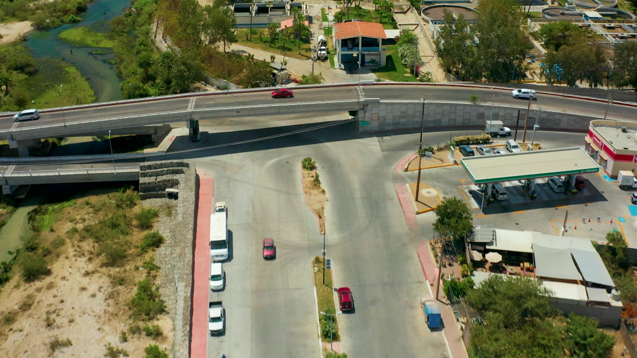 Aerial frame from San Jose del Cabo showing a bridge over a green-edged river, cars on the overpass, a gas station below, parked vehicles, utility tanks, and no visible people.