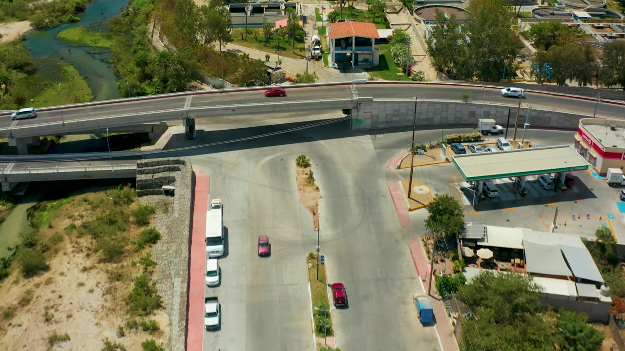 Aerial view in San Jose del Cabo with the bridge centered, a red car and white car on the overpass, the gas station canopy below, river water at upper left, and no visible people.