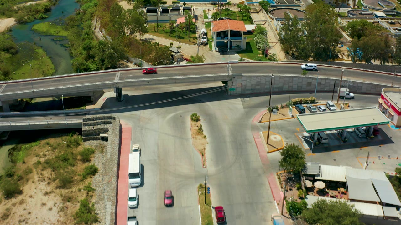 San Jose del Cabo aerial frame showing the bridge stretching across the image, a white car on the road, gas pumps under a canopy, parked cars, industrial tanks, river greenery, and no visible people.
