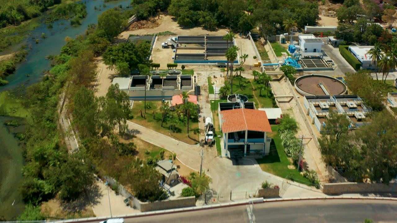 Aerial view in San Jose del Cabo showing a water treatment plant with rectangular basins, a circular clarifier, service roads, trees, green river water, and no visible people.