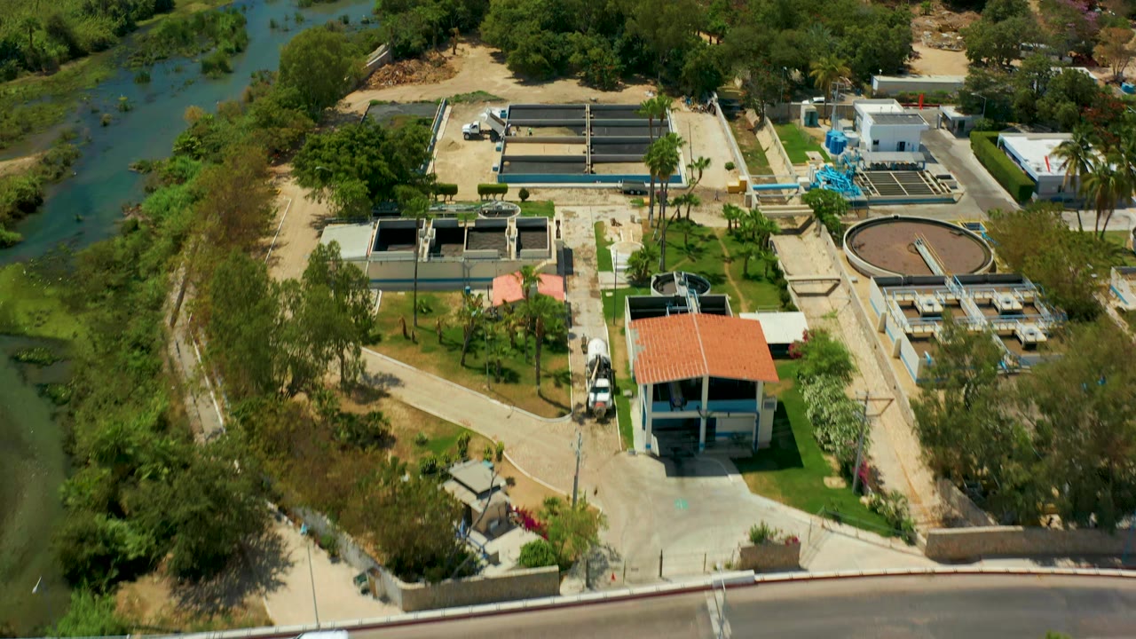 Elevated San Jose del Cabo view of concrete treatment tanks, a round clarifier, blue piping or equipment, a red roofed structure, roadside pavement, river water, trees, and no visible people.