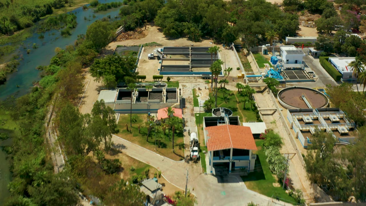 High aerial frame in San Jose del Cabo with the treatment plant, circular clarifier, rectangular basins, white service buildings, blue equipment, riverbank greenery, and no visible people.