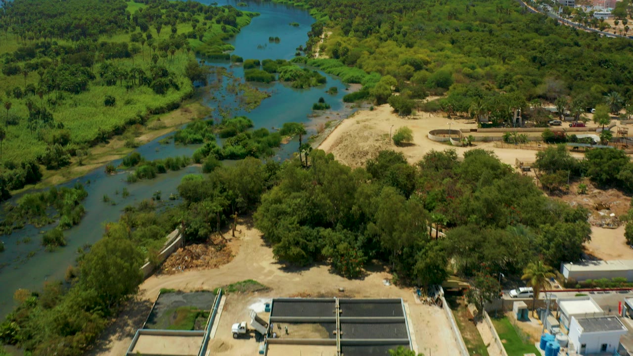 Early aerial frame over San Jose del Cabo showing a blue-green river through dense vegetation, sandy ground, small structures near the lower edge, and no visible people.