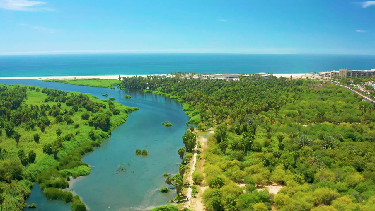 Wide aerial frame in San Jose del Cabo with the river curving through green brush toward a sandy beach and blue ocean, with no visible people.
