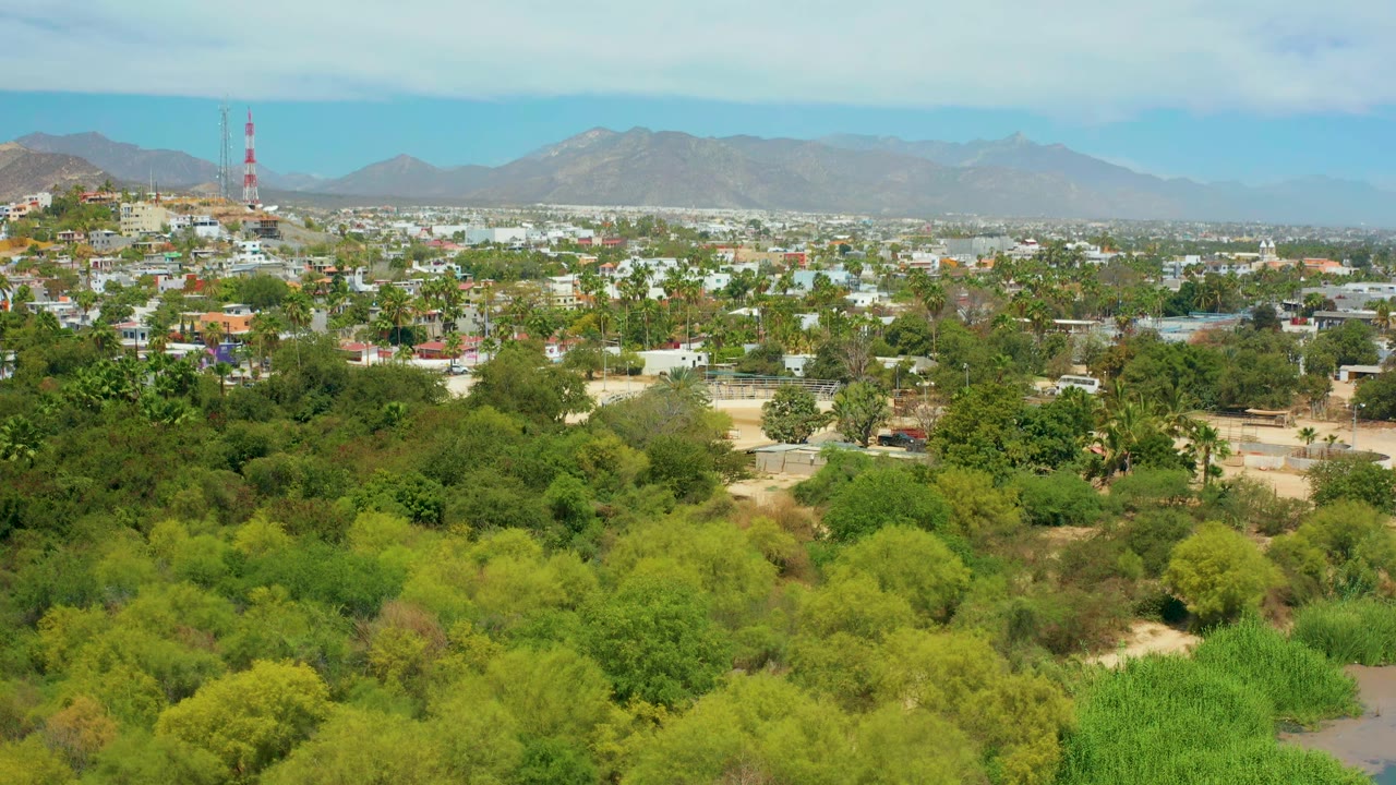 Early runtime view of San Jose del Cabo showing layered green treetops, scattered low buildings, a red and white tower, mountains, pale clouds, and no visible people.