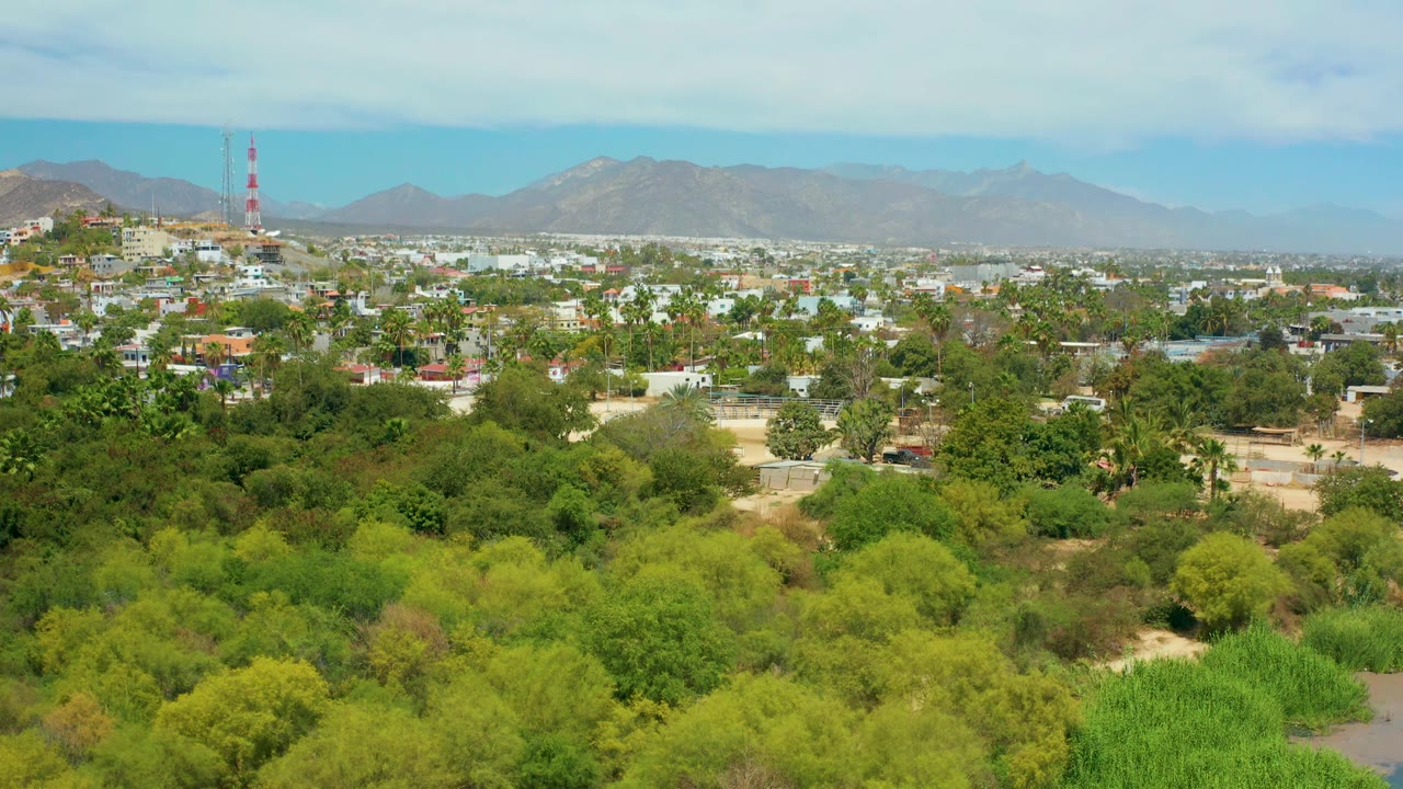 Aerial frame over San Jose del Cabo with bright green foreground vegetation, white and tan buildings, hazy mountains, a partly cloudy sky, and no visible people.
