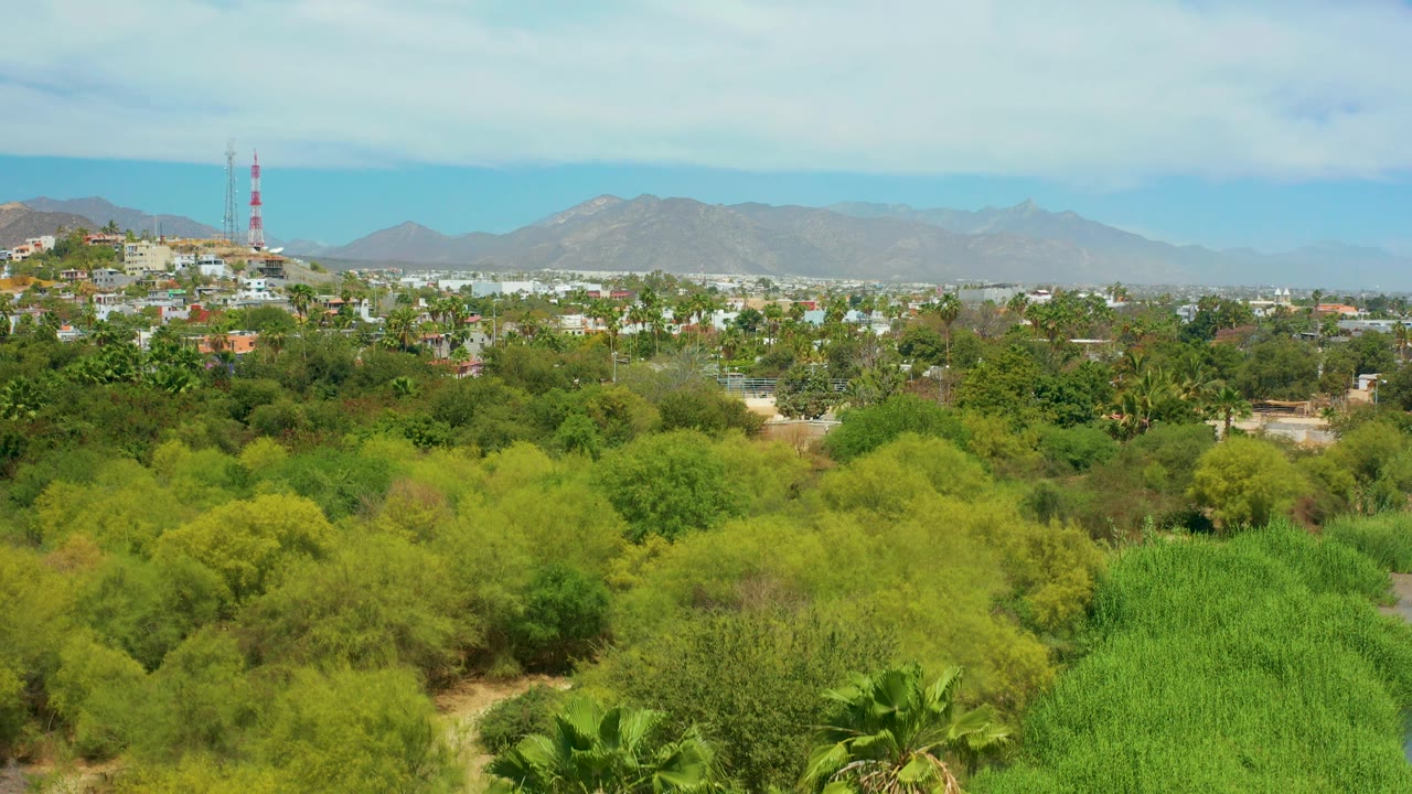 A later frame shows San Jose del Cabo's palm canopy and shrubs below a strip of buildings, with hazy gray mountains and broken cloud cover above, and no visible people.