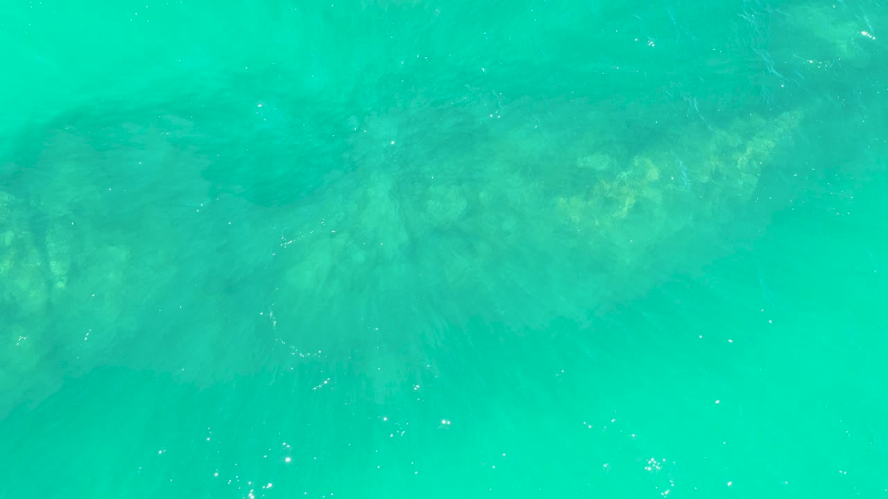 Calm turquoise ocean water at Marquis Los Cabos covers a rocky bottom with a few white bubbles crossing the lower area and no visible people.