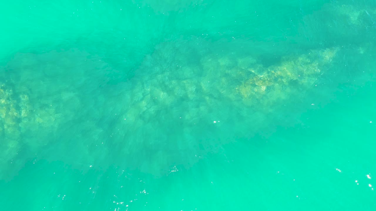 Overhead turquoise ocean water at Marquis Los Cabos shows the submerged reef band slightly left of center with small bright surface flecks and no visible people.