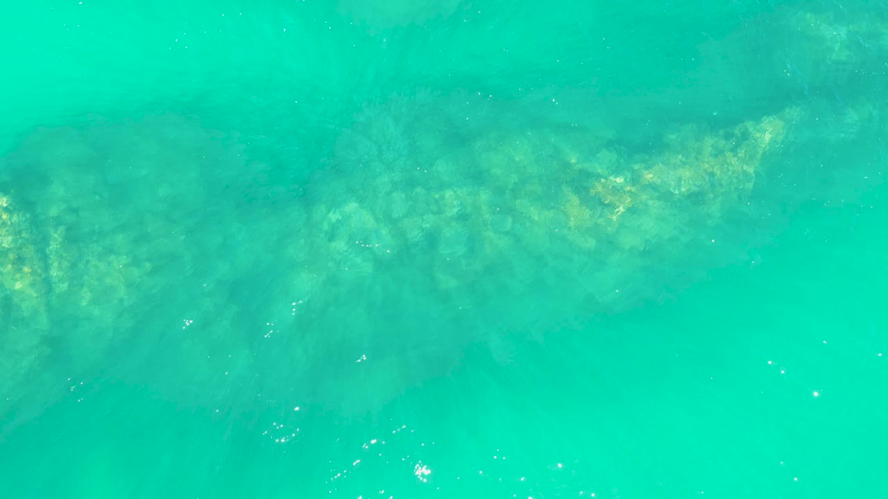 A darker submerged reef form sits beneath bright turquoise water at Marquis Los Cabos, with a few white bubbles on the surface and no visible people.