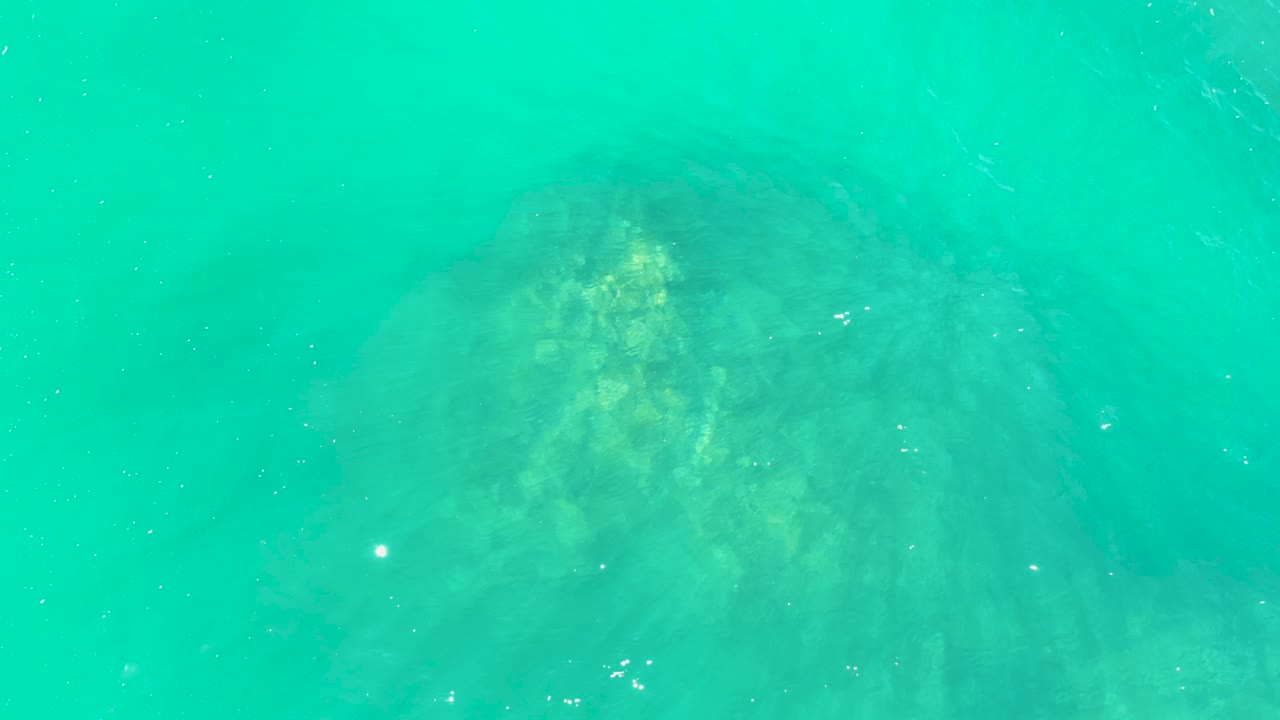 Clear turquoise water at Marquis Los Cabos reveals the rock formation centered lower in the frame, with faint particles and no visible people.