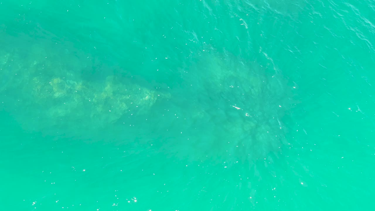 A high overhead frame shows turquoise ocean water at Marquis Los Cabos with yellow-green patches on the submerged structure and no visible people.