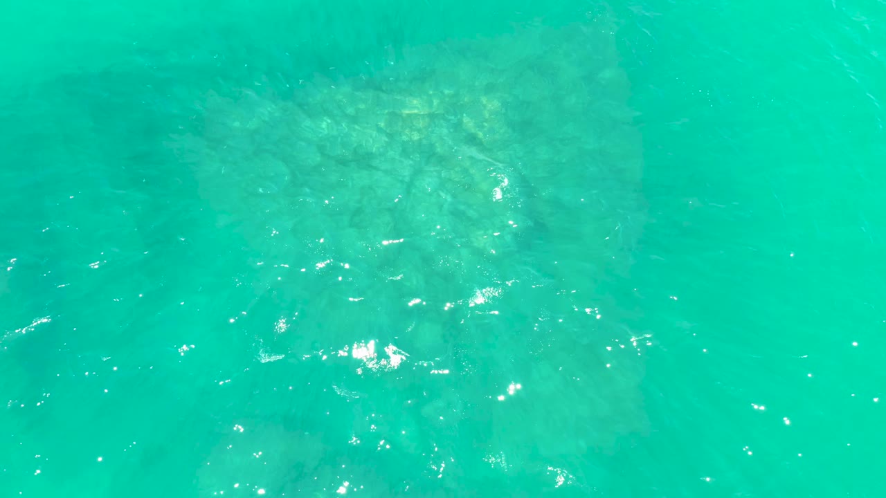 Turquoise ocean water at Marquis Los Cabos fills the frame, with the submerged formation showing more centrally beneath gentle ripples and no visible people.
