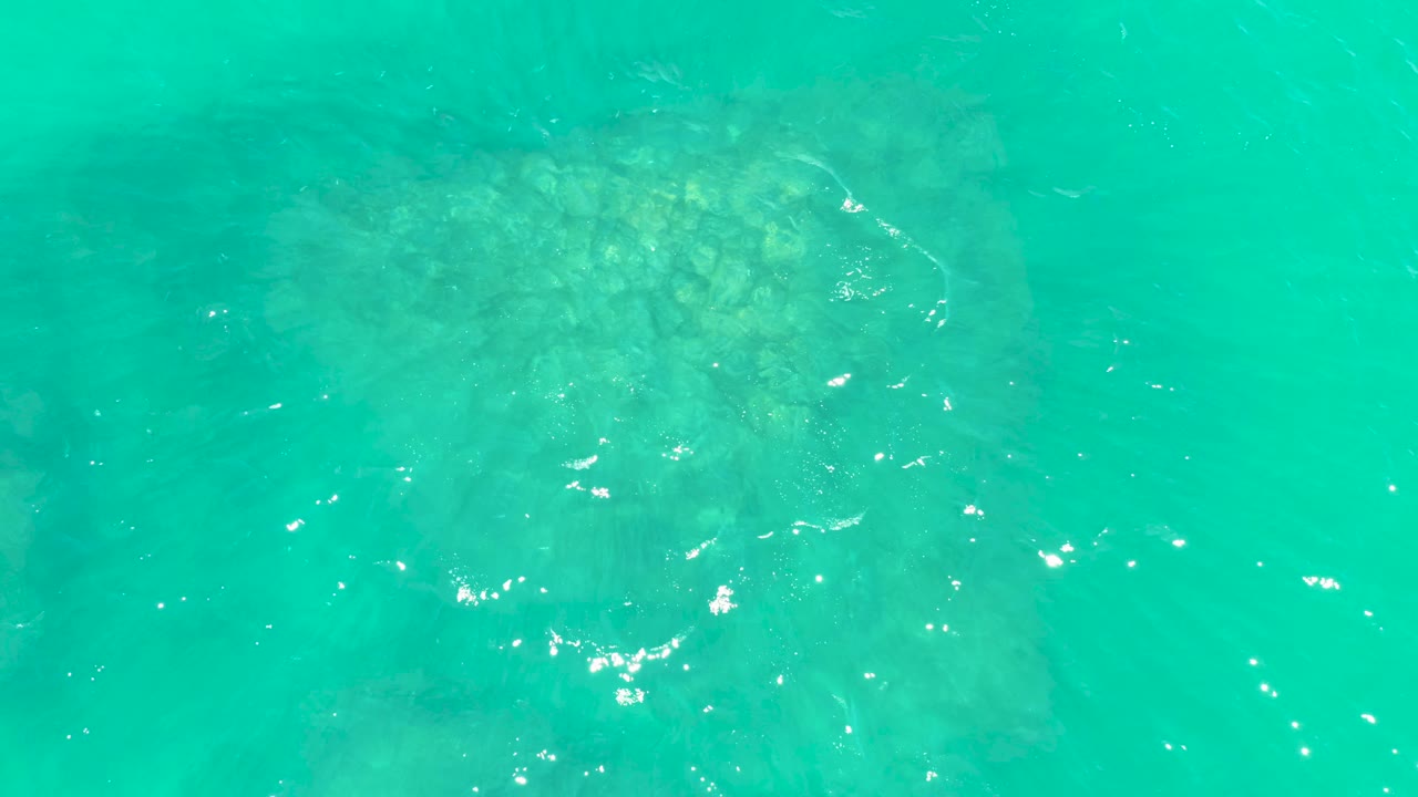 A top-down turquoise water frame at Marquis Los Cabos shows the submerged formation toward the upper right, with small white reflections across the surface and no visible people.