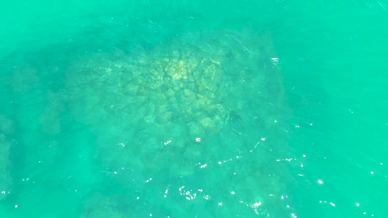 Clear turquoise water at Marquis Los Cabos reveals a darker submerged rock area in the lower left, with a brighter yellow-green patch nearby and no visible people.