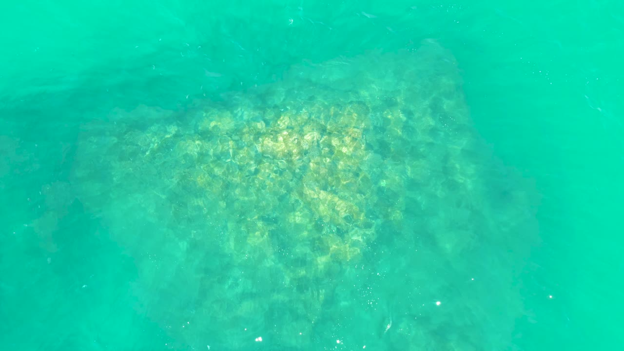 A near-vertical underwater view at Marquis Los Cabos shows the rock formation centered under clear turquoise water with small suspended particles and no visible people.