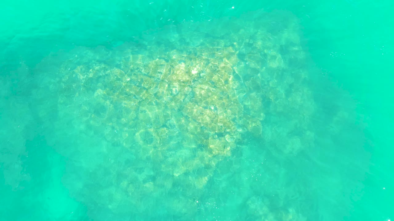 At 0:03, the sea turtle is partly softened by surface ripples in clear turquoise water at Marquis Los Cabos, with rocky seabed texture visible around it and no visible people.