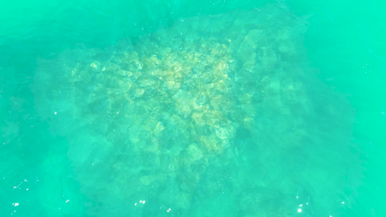 Clear blue-green water reveals a central patch of rocks on the ocean floor at Marquis Los Cabos, with small highlights on the surface and no visible people.